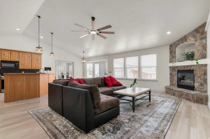 Spacious living area featuring vaulted ceilings, a stone-clad fireplace with hearth, multiple windows, wood-finish flooring, and a kitchen bar with wood cabinetry and black tile backsplash