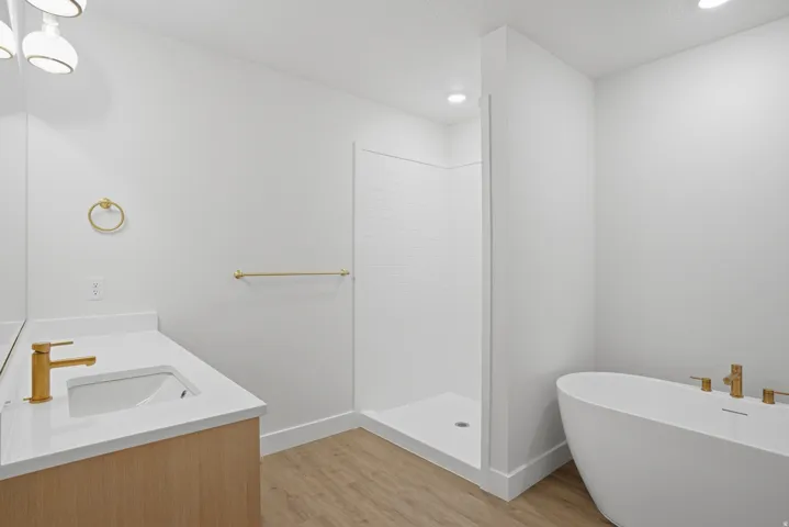 White freestanding soaking tub, walk-in shower with subway tile, wood-finish flooring, single vanity with white countertop, and gold-finish fixtures