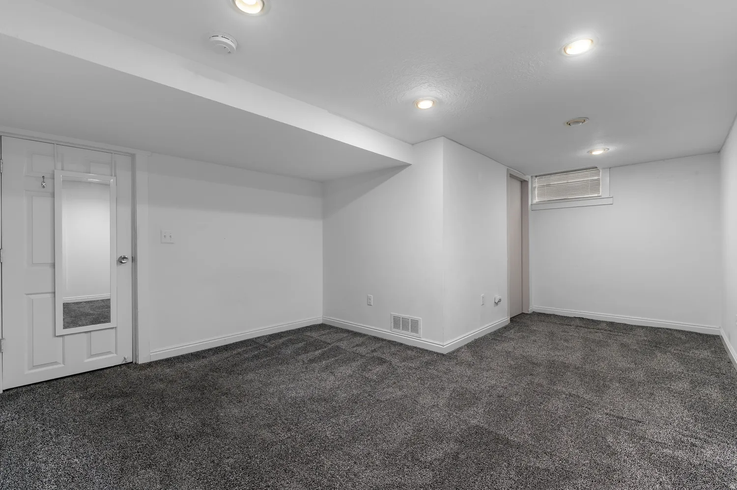 Recessed lighting, gray carpeting, white walls, and a window provide illumination