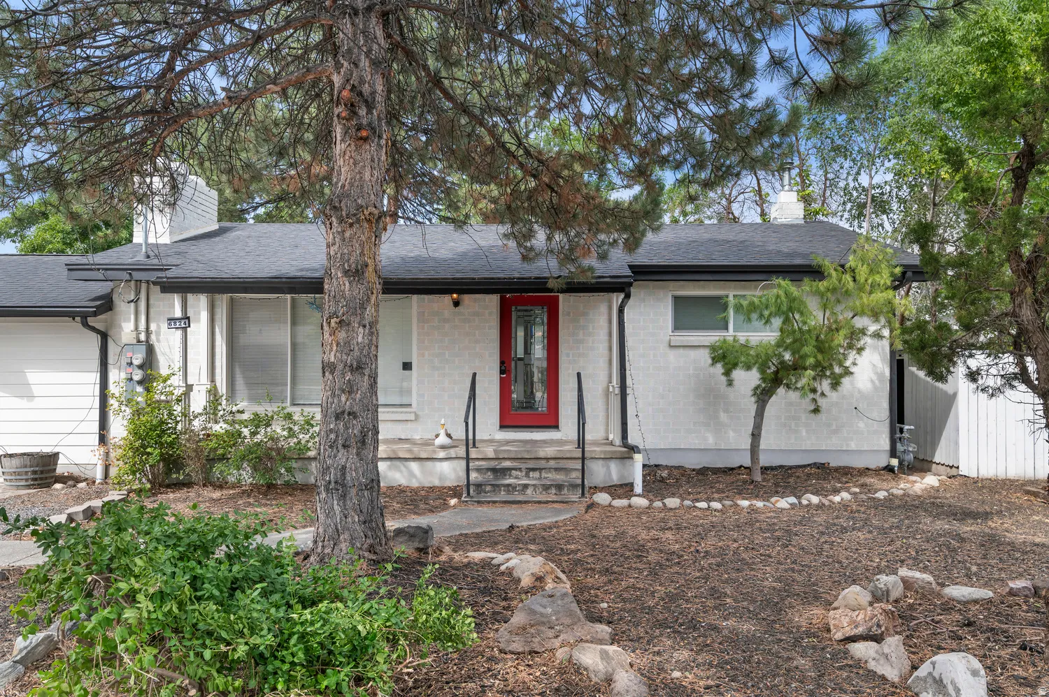 White brick exterior featuring a contrasting red front door, dark shingle roof, and a stone-edged garden bed