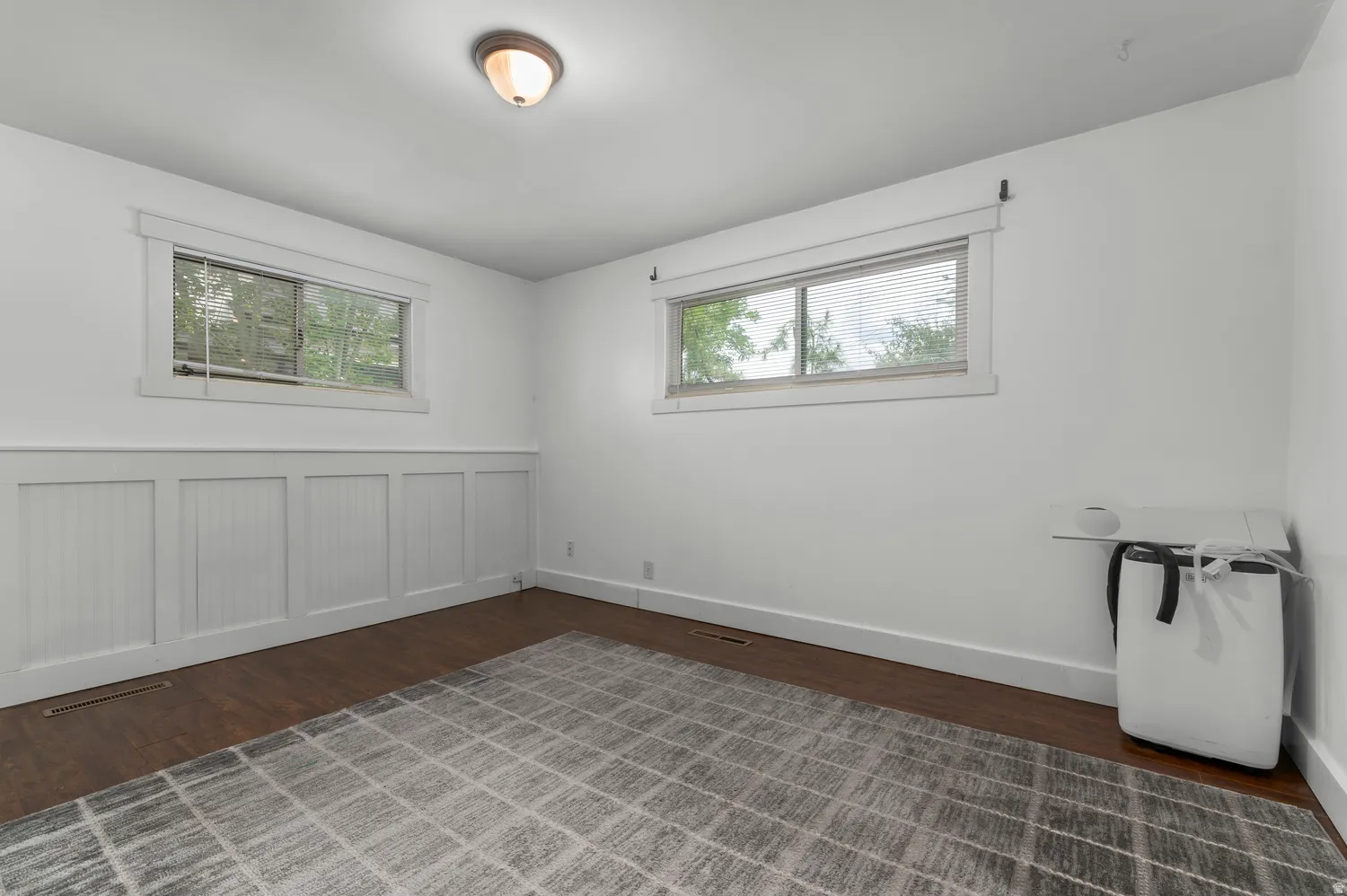 Dark wood-finish flooring, white board and batten wall accent, two distinct windows, and a flush-mount ceiling light fixture