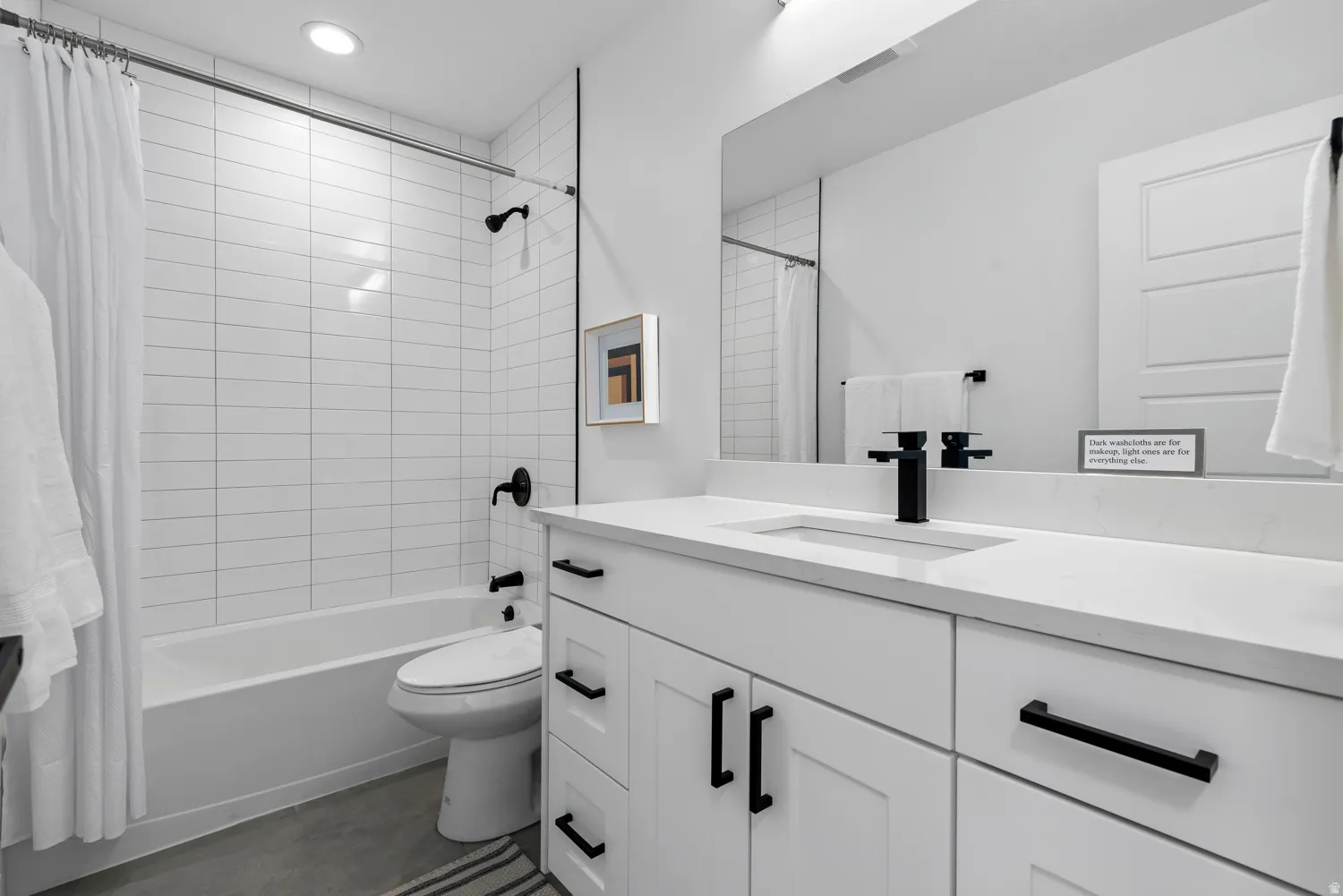 Unit #2White tiled shower-tub combination, single vanity with integrated sink, stone countertop, matte black hardware, and gray concrete-finish flooring