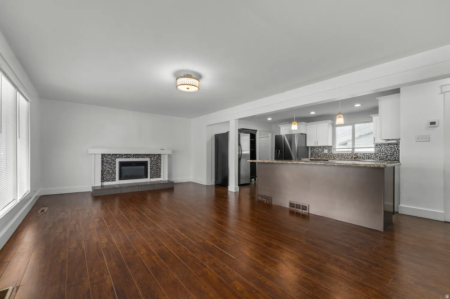 Wood-finish flooring throughout, a modern fireplace with a tiled surround, a kitchen island with pendant lighting, white cabinetry, and stainless steel appliances