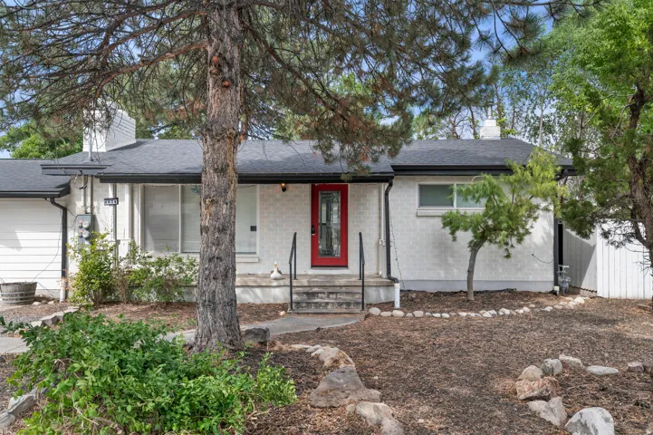 White brick exterior featuring a contrasting red front door, dark shingle roof, and a stone-edged garden bed