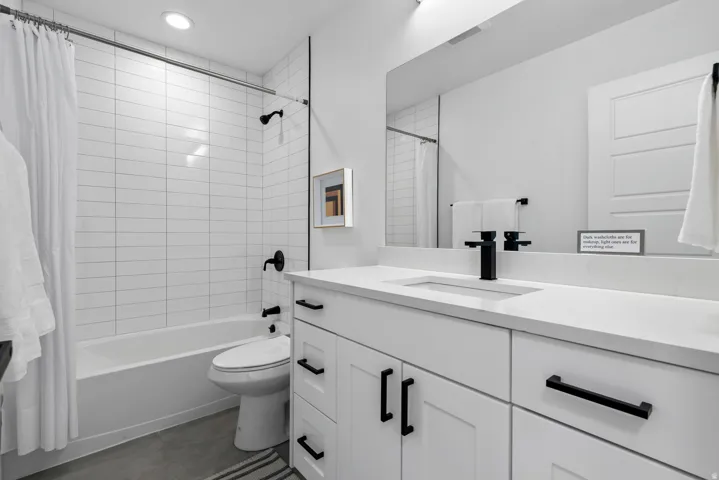 Unit #2White tiled shower-tub combination, single vanity with integrated sink, stone countertop, matte black hardware, and gray concrete-finish flooring