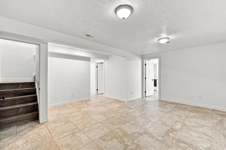 Spacious room featuring neutral tile flooring, white walls, overhead flush-mount lighting, and wood-finish stairs