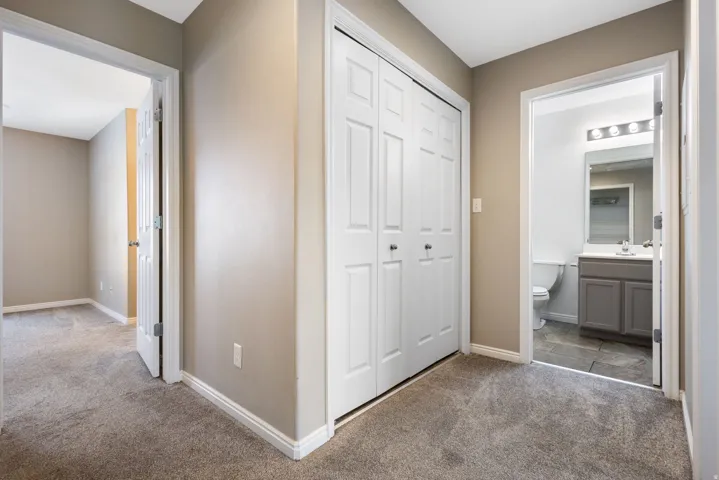 Carpeted hallway with a bi-fold closet, leading to a room with carpet flooring and a bathroom with tile flooring, a gray vanity, and a mirror with sconce lighting
