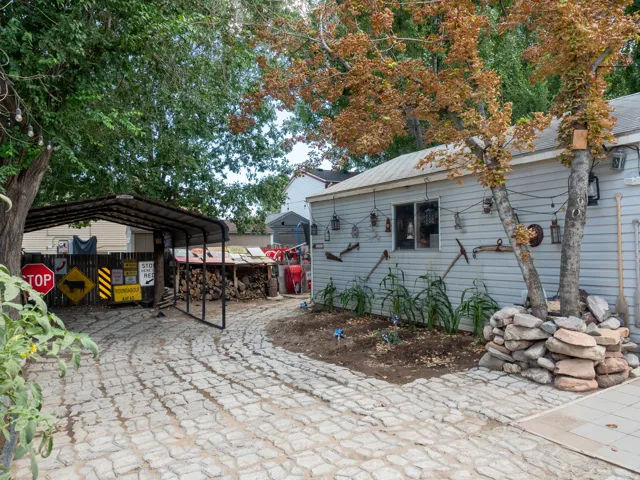 Textured paver driveway, carport structure, wood siding on exterior, established trees, and stone landscaping