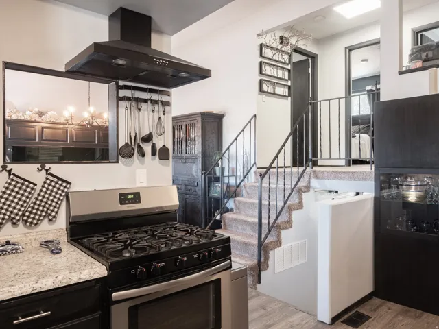 Stainless steel gas range with a black overhead range hood, light-toned countertop, dark wood-finish cabinetry, and a carpeted staircase with wrought iron railings