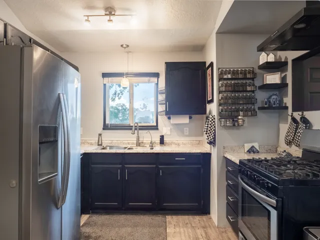 Stainless steel side-by-side refrigerator, stainless steel sink with gooseneck faucet, dark cabinetry, light-toned countertops, and wood-finish flooring