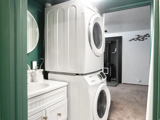 White stacked laundry appliances, white vanity with integrated sink, circular wall mirror, and green wall paneling