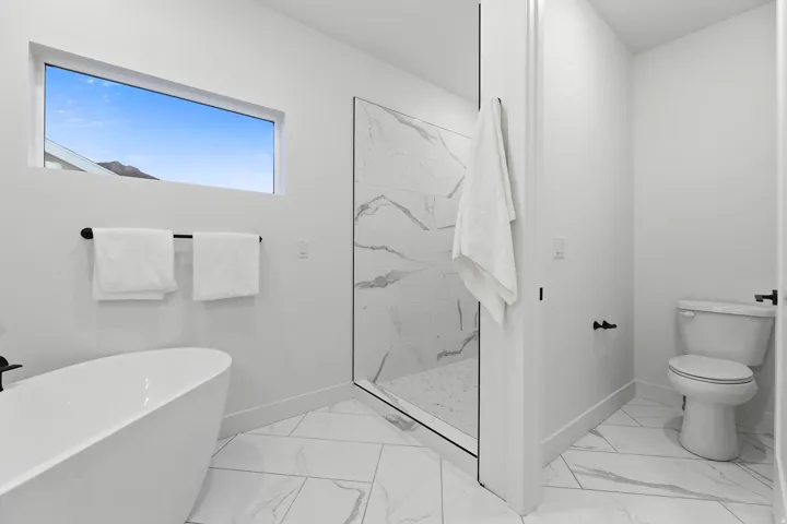 Freestanding soaking tub, walk-in shower with glass enclosure, white tile flooring, and a rectangular transom window