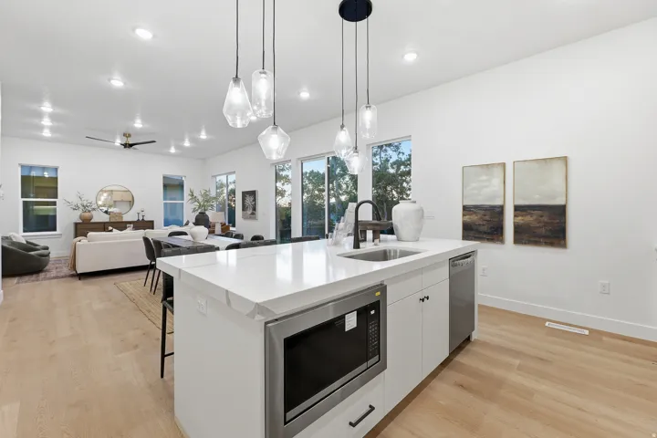 Spacious open-concept living area featuring wood-finish flooring, a kitchen island with integrated sink and microwave, pendant lighting, and abundant natural light from multiple windows