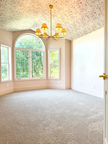 Expansive room featuring a large arched window, multiple rectangular windows, patterned ceiling wallpaper, a brass chandelier with six shaded lights, and textured carpet flooring