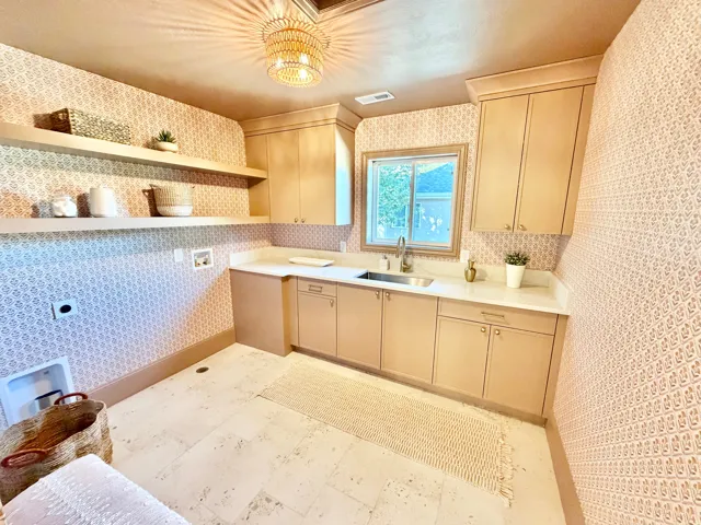 Neutral-toned cabinetry with brass hardware, undermount stainless steel sink, light-toned countertops, and decorative wall covering
