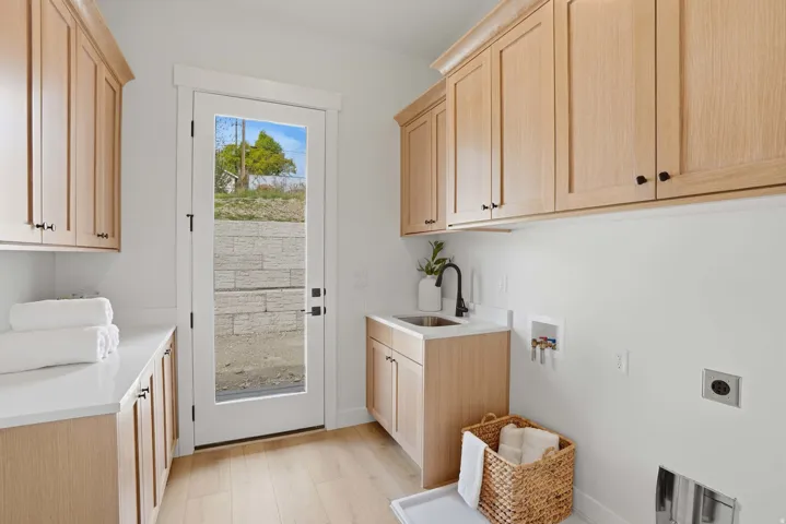 Light wood-finish cabinetry, white solid surface countertops, wood-finish flooring, a utility sink with a matte black faucet, and a glass-paned door