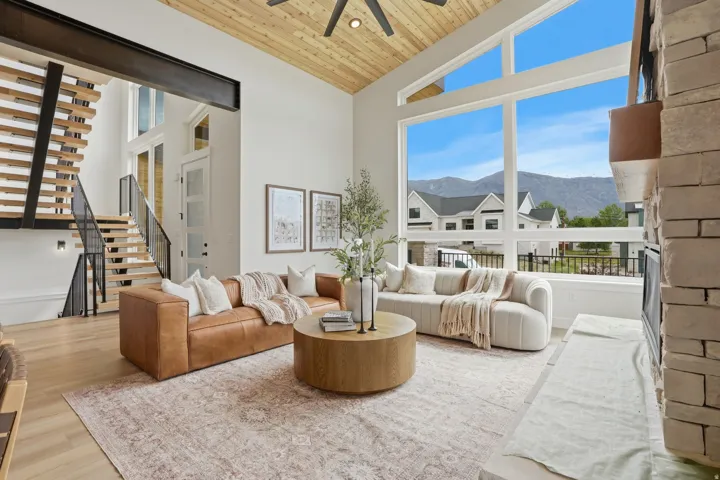 Vaulted wood-finish ceiling, expansive windows, light wood-finish flooring, stone-clad fireplace, and an open-tread staircase with metal railings