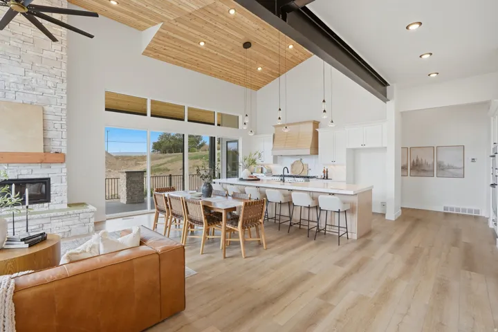 Vaulted ceiling with wood-finish accents, stone-faced fireplace, large sliding glass doors, center island with seating, and wood-finish flooring