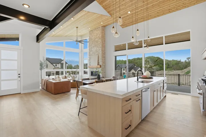 Vaulted wood-paneled ceiling with exposed beams, full-height stone fireplace, wood-finish flooring, large multi-pane windows, and a kitchen island with built-in cabinetry