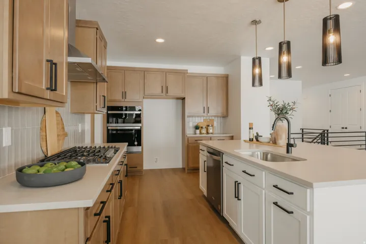 Open-concept kitchen featuring wood-finish flooring, light wood cabinetry, a stainless steel range hood, and a kitchen island with a built-in sink and pendant lighting