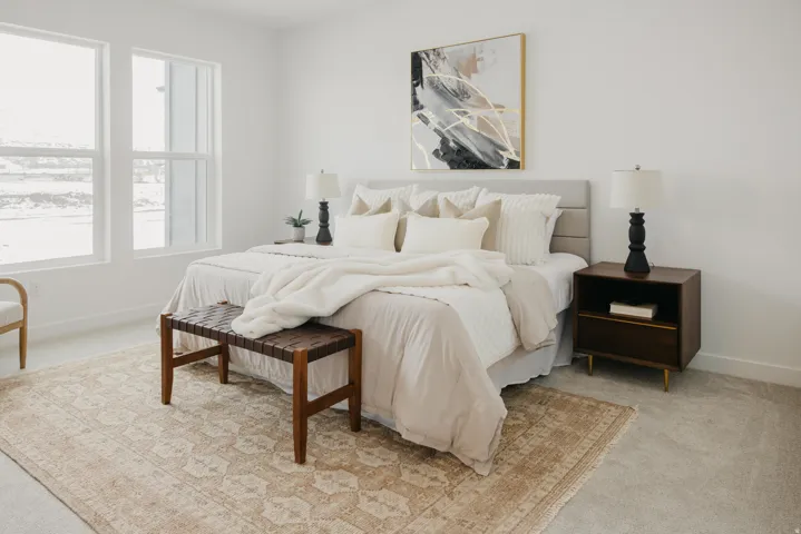 Bright bedroom featuring two large windows, carpet flooring, a built-in upholstered headboard, and a wood-finish nightstand