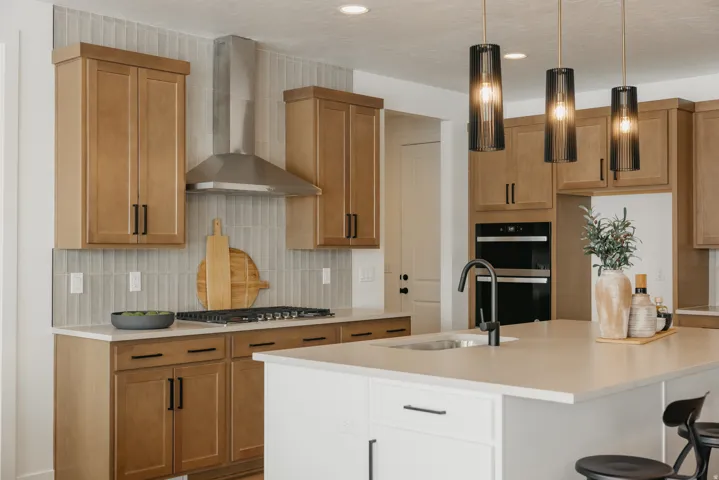 Kitchen featuring light wood cabinetry, a stainless steel range hood, built-in double ovens, a center island with sink, and pendant lighting