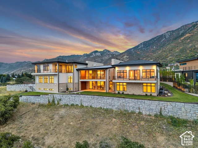 Contemporary architectural style featuring expansive glazing, multiple balconies with glass railings, a stone retaining wall, and mountain scenery