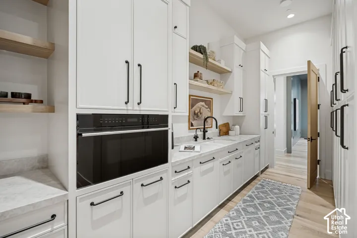 White cabinetry with black hardware, built-in oven, integrated sink with matte black faucet, light-toned wood-finish flooring, and open wood shelving