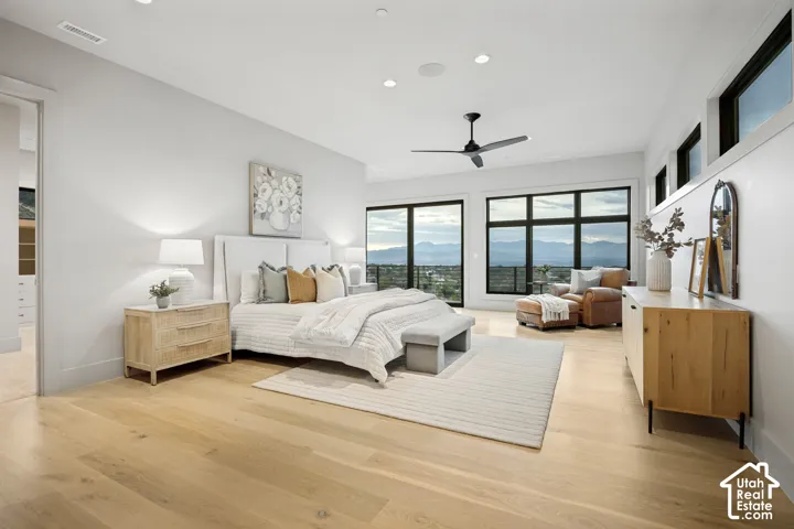 Expansive light wood-finish flooring, large windows with black frames, modern ceiling fan, recessed lighting, and a white upholstered headboard