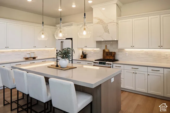 Spacious kitchen featuring white cabinetry, light-toned countertops, a large center island, wood-finish flooring, and a range with a decorative hood