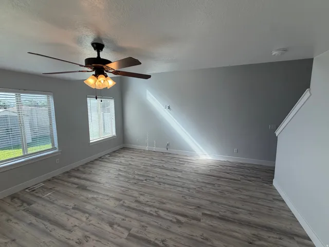Spacious room featuring wood-finish flooring, multiple windows with blinds, a ceiling fan with integrated lighting, and neutral wall paint