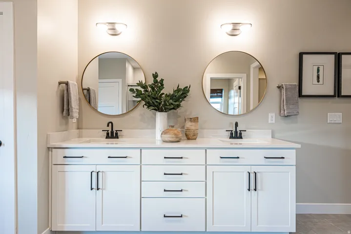 Dual vanity with white cabinetry and matte black hardware, stone countertop, two round mirrors, and wall-mounted light fixtures