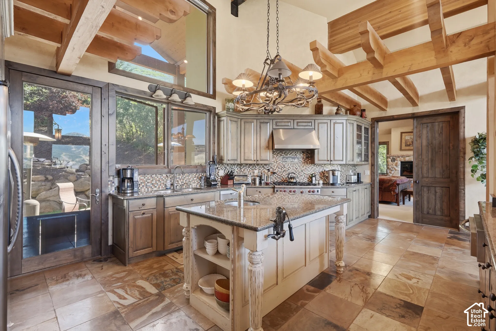 Exposed wood beam ceiling, full tile flooring, center island with integrated shelving, patterned tile backsplash, and stainless steel range hood