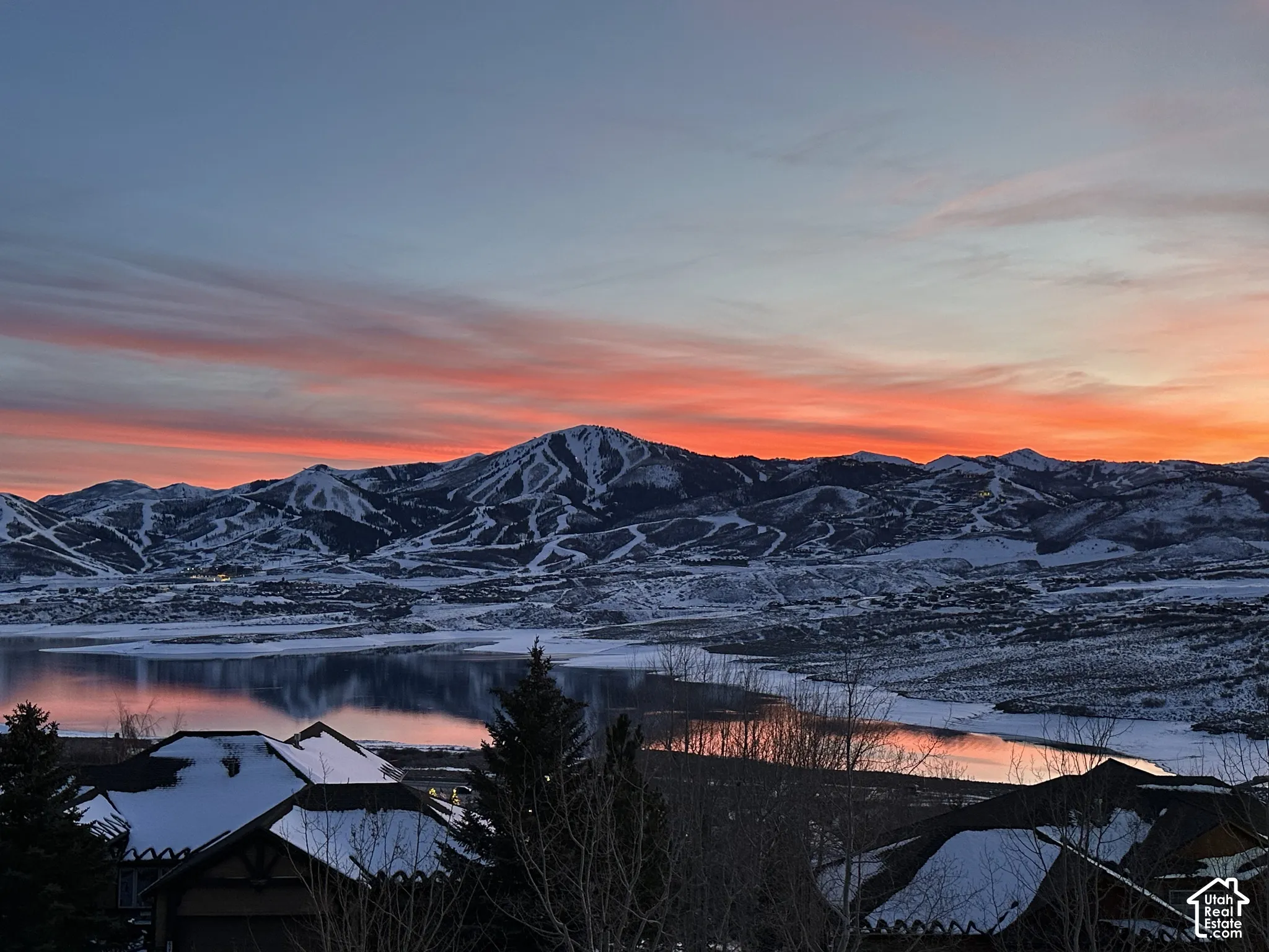 Snow-capped mountain range with visible ski slopes, large body of water, and residential structures with snow-covered roofs
