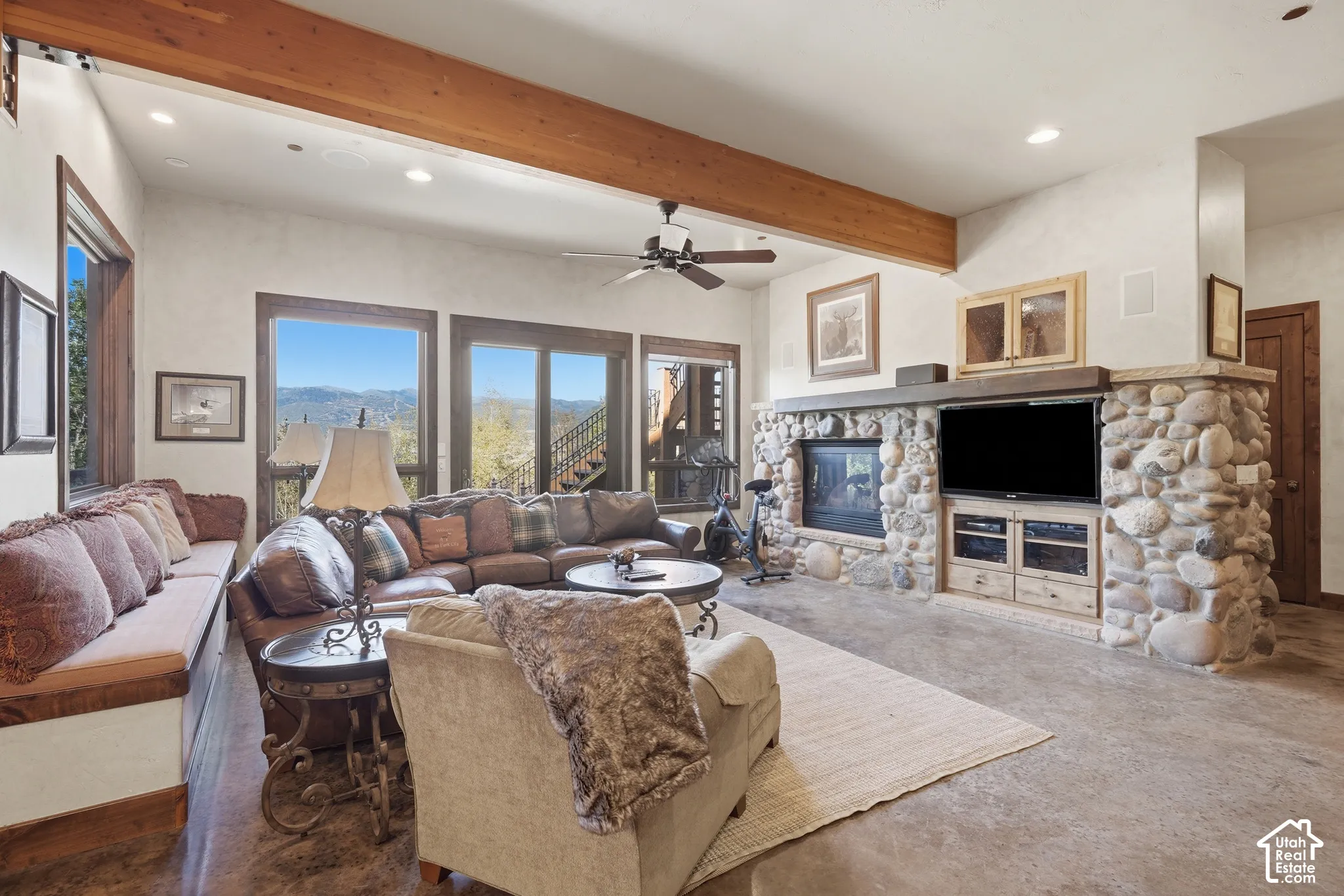 Exposed wooden beam, stone fireplace with hearth, wood-frame windows, built-in window seating, and a ceiling fan