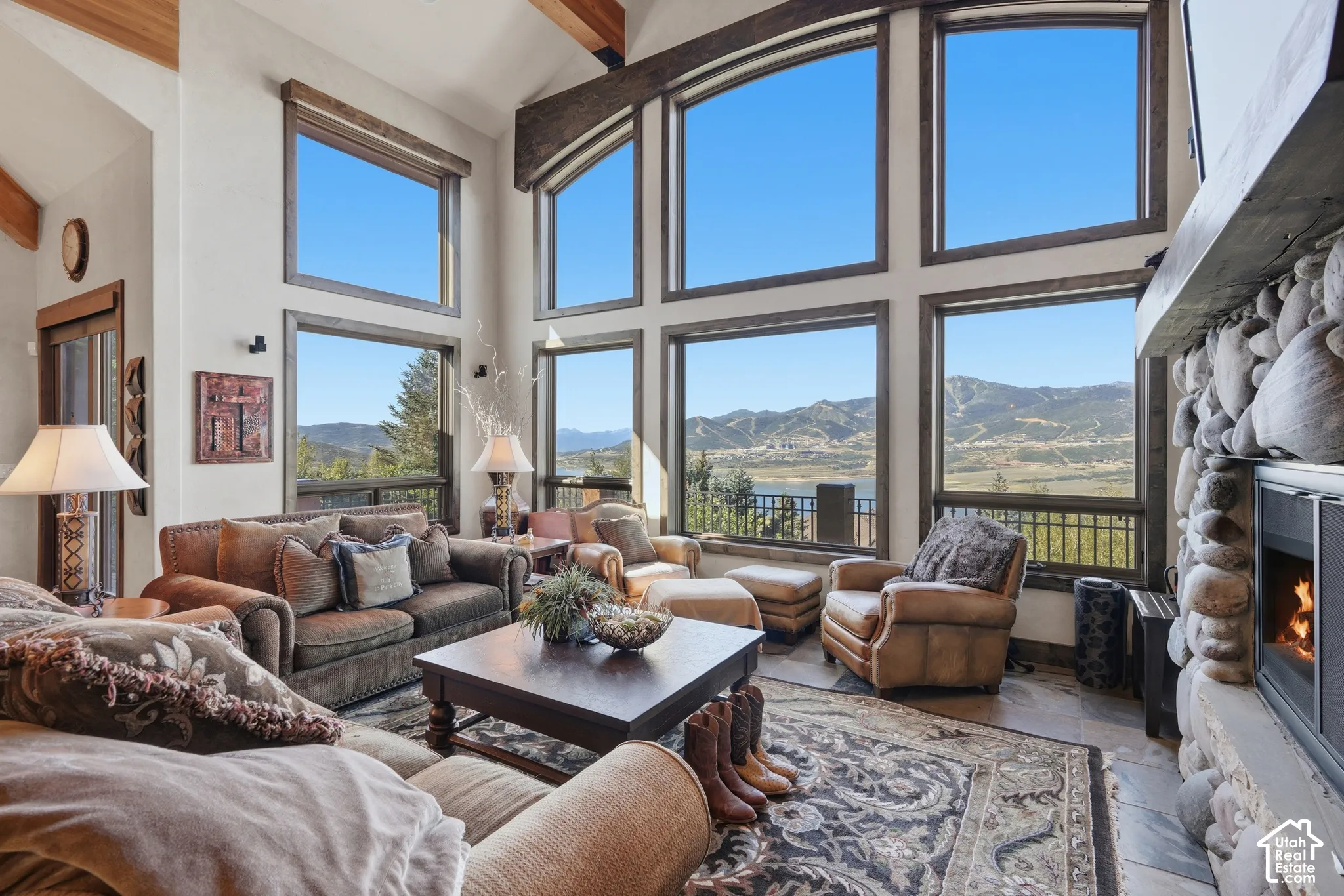 Spacious living area featuring a stone-clad fireplace, soaring ceiling, exposed wood beams, and expansive windows showcasing a mountain and lake landscape