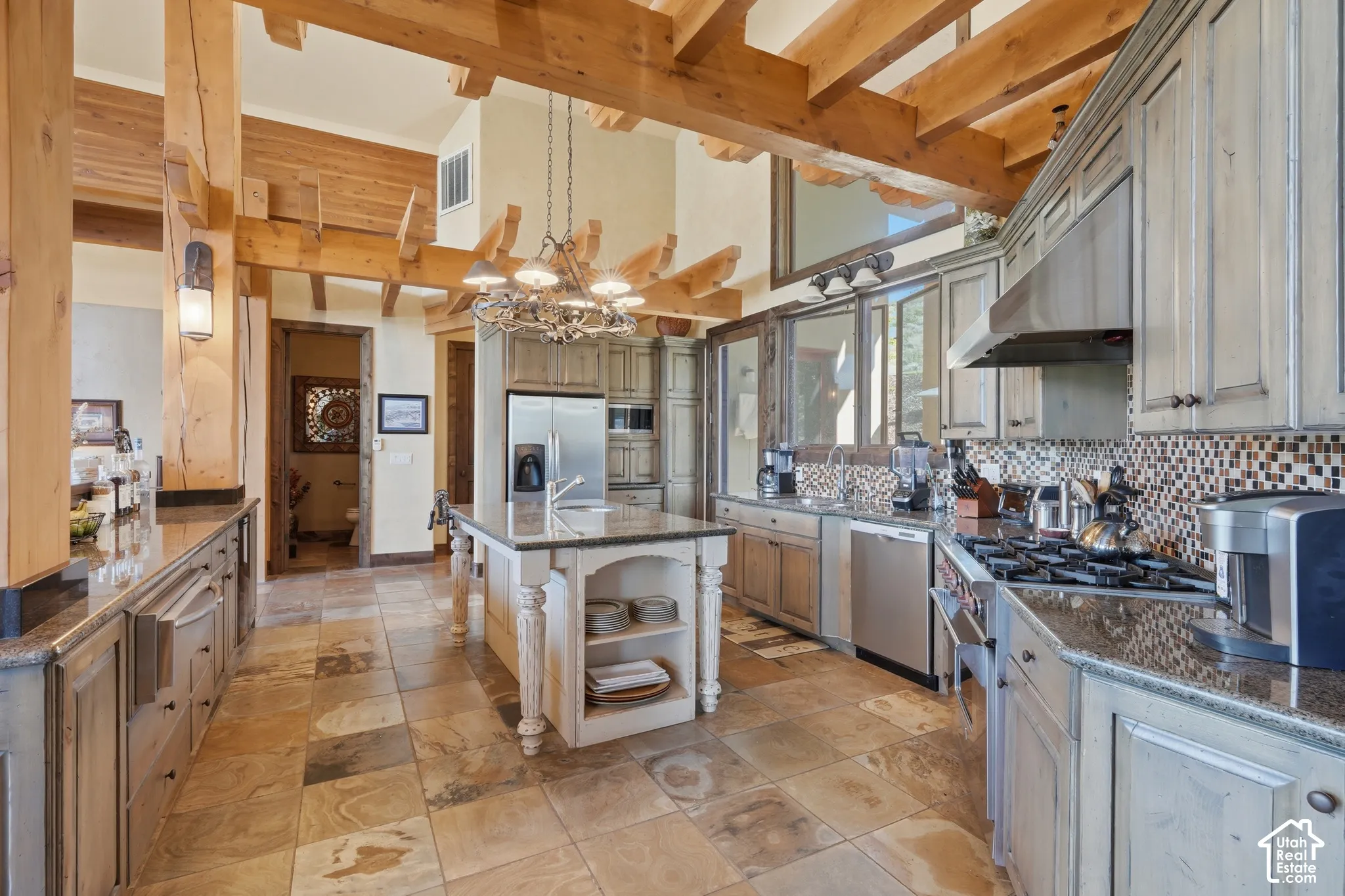 Kitchen featuring exposed wood beams, stone tile flooring, a central island with an integrated sink, granite countertops, and stainless steel appliances