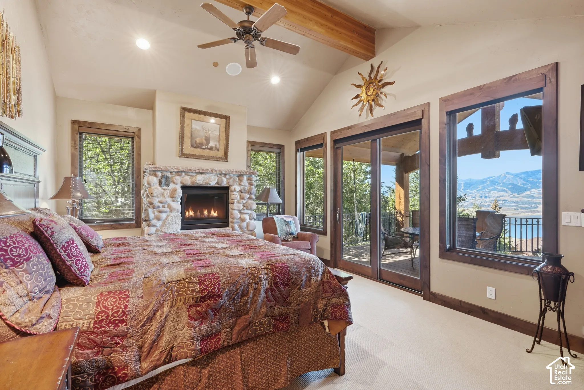 Stone-clad fireplace with a built-in firebox, vaulted ceiling with exposed wood beams, ceiling fan with integrated lighting, wood-trimmed windows, and sliding glass doors accessing an outdoor area