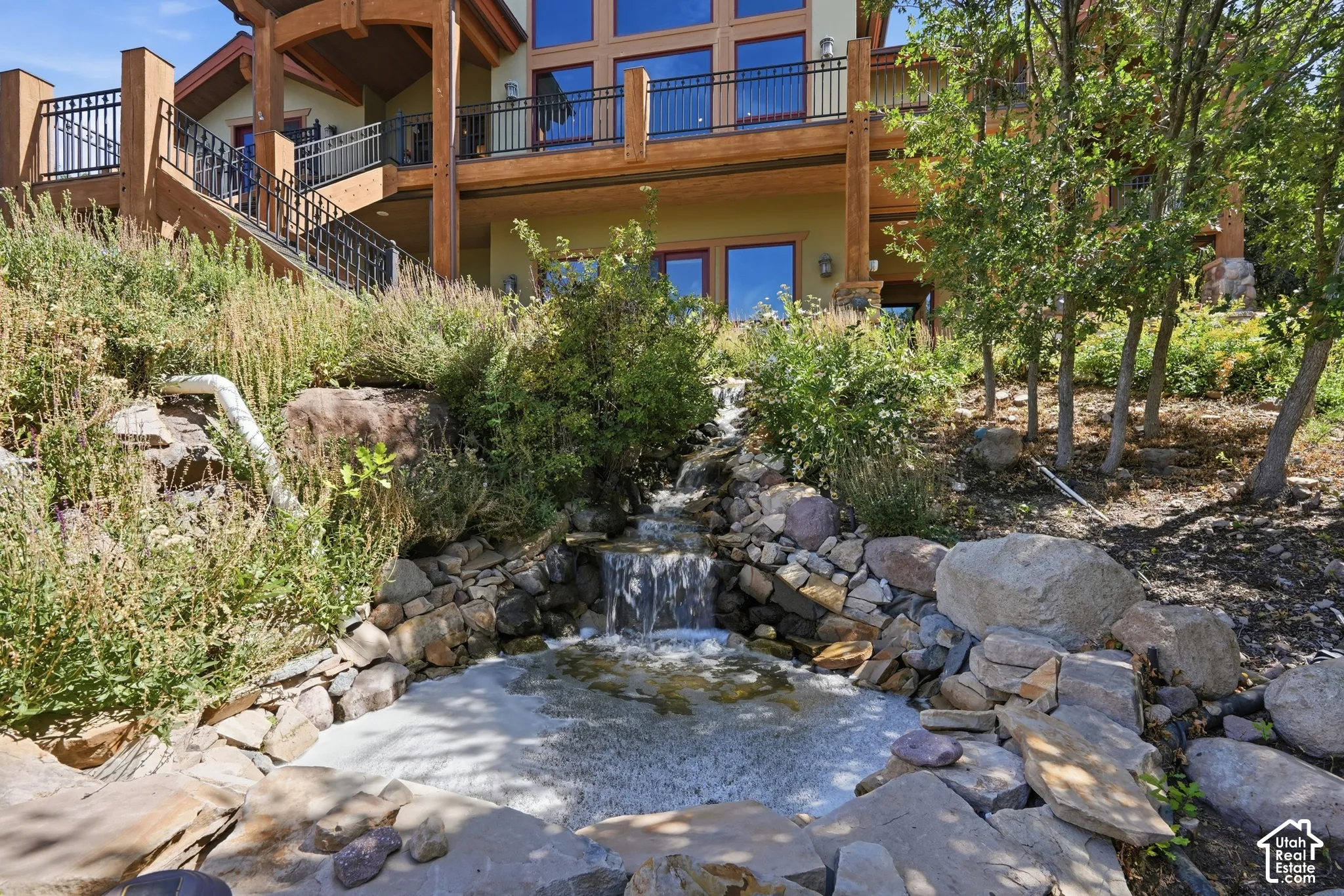 Multi-level residence with expansive balconies and exterior staircase, featuring a rock-lined water feature and established landscaping
