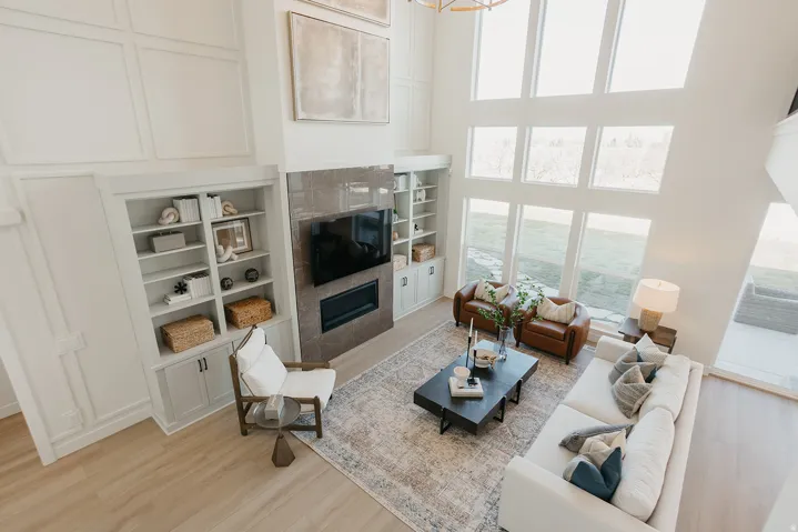 Expansive living area featuring a two-story ceiling, a fireplace with a stone surround, built-in shelving, large picture windows, and wood-finish flooring