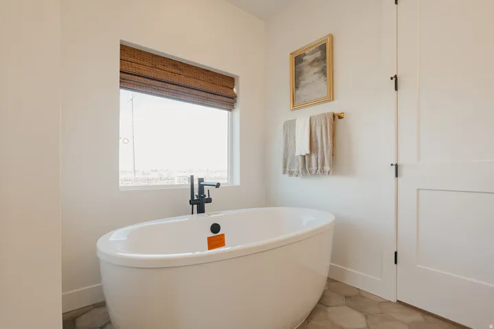Freestanding soaking tub with a matte black faucet, hexagonal floor tiling, and a window with a woven shade