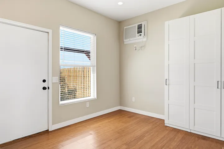 Warm wood-finish flooring, built-in white cabinetry, window with blinds, recessed lighting, and wall-mounted air conditioning unit