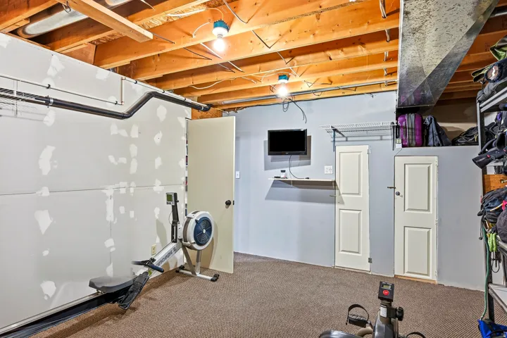 Unfinished basement space with exposed wood joists, carpeted flooring, and a drywall partition with a door