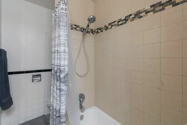 Bathtub with a shower fixture, light-toned wall tiling, and decorative mosaic accent band