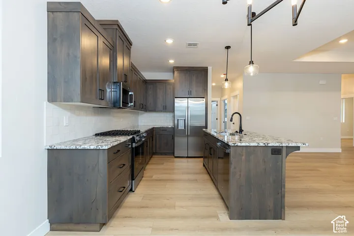 Kitchen featuring wood-finish cabinetry, light-toned countertops, a full suite of stainless steel appliances, a tile backsplash, and an island with a sink and pendant lighting