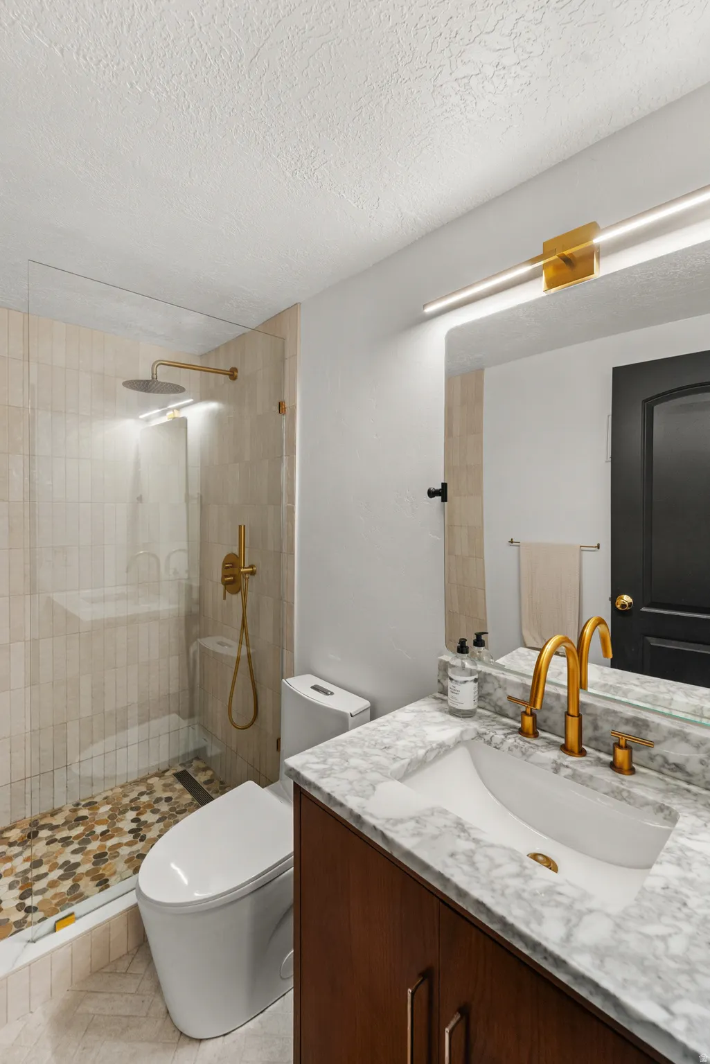 Glass-enclosed shower with a rain shower head and handheld sprayer, vanity with a marble countertop, undermount sink, and gold-finish fixtures, wood-finish cabinetry, and mosaic tile shower floor