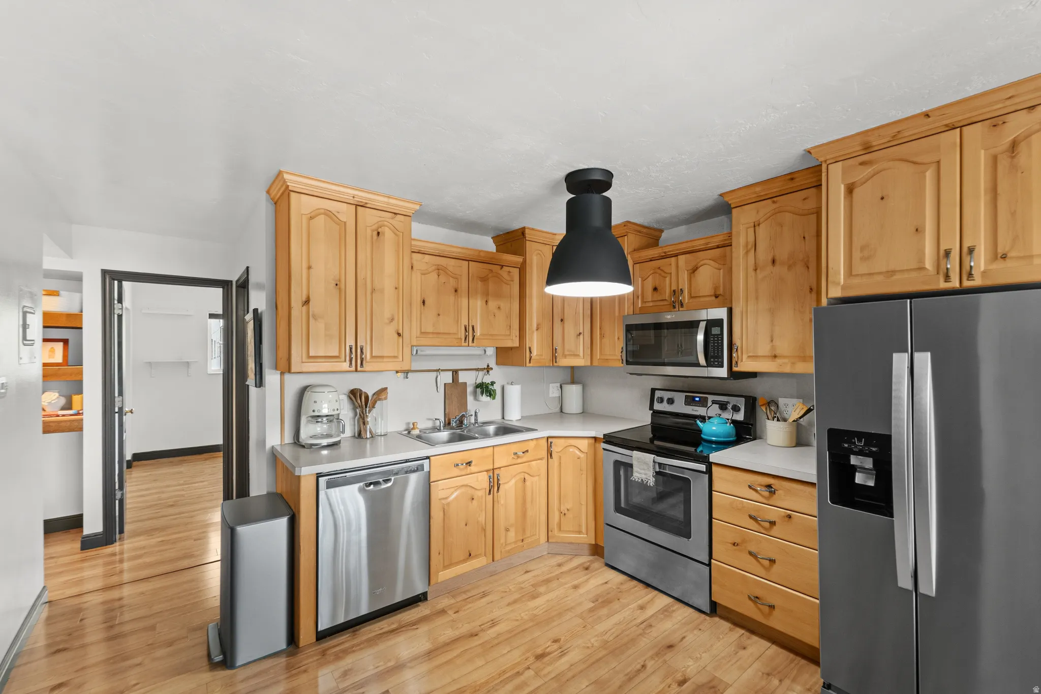 Wood-finish flooring, light wood cabinetry, stainless steel appliances, white countertops, and a black pendant light