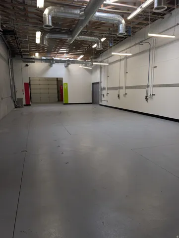 Exposed wood ceiling beams, overhead ductwork, multiple linear lighting fixtures, concrete flooring, and a roll-up door