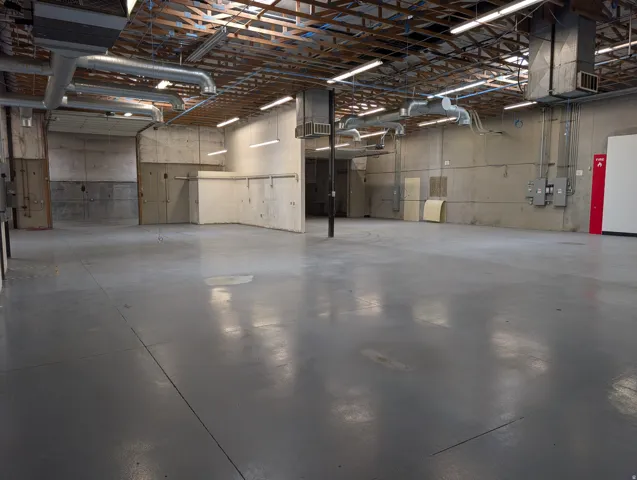 Expansive open-concept space featuring concrete walls, exposed wood beam ceiling, overhead ductwork, and fluorescent lighting fixtures