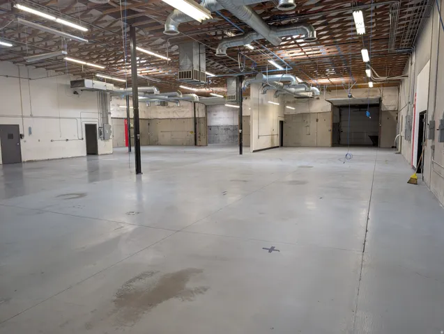 Large open warehouse space featuring a high ceiling, visible ductwork, overhead fluorescent lighting, concrete flooring, and an overhead door